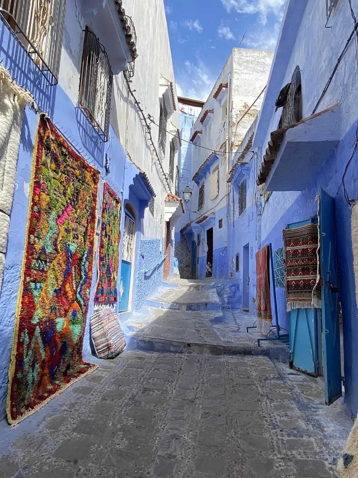Ruelle en pente de la médina : la partie basse des murs est peinte en bleu, les niveaux supérieurs sont blancs, de grands tapis berbères multicolores pendent au mur de gauche, des tapis à franges sont présentés sur support à droite, et le sol est un dallage de pierre en marches
