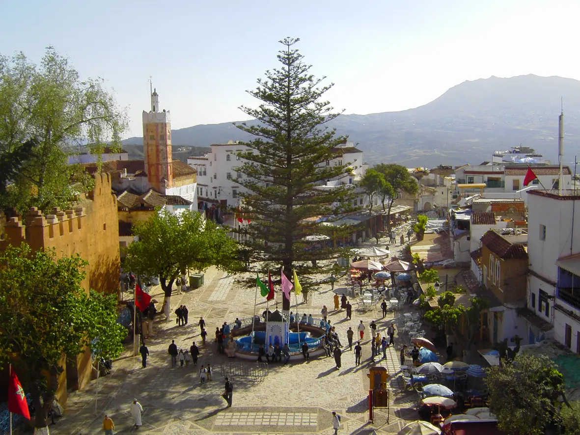 Outa el Hammam prise d'en haut : les remparts ocre et crénelés de la kasbah à gauche, un très grand araucaria au centre, la fontaine au carrelage bleu entourée de quatre fanions colorés, et la place en pleine activité au soleil, entre les cafés