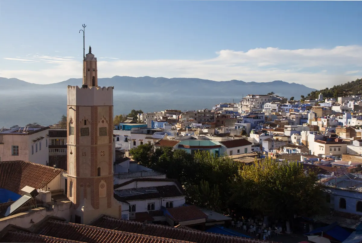 Vue plongeante sur les toits de Chefchaouen : à gauche, au premier plan, le minaret de brique de la Grande Mosquée, les toits blancs et leurs tuiles rouges qui s'étalent vers la droite, et la crête du Rif dans la brume au fond