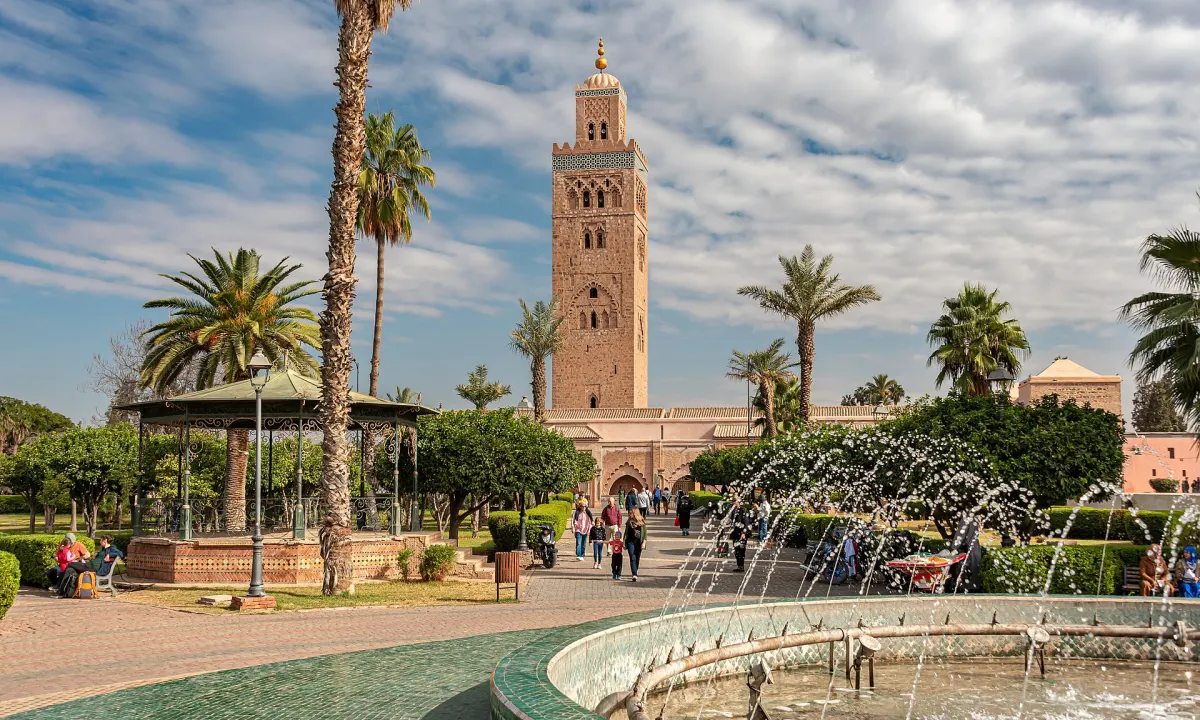 Le minaret de la Koutoubia dominant le parc qui l’entoure, entre les palmiers, avec un kiosque à toit vert et le jet d’eau de la fontaine au premier plan