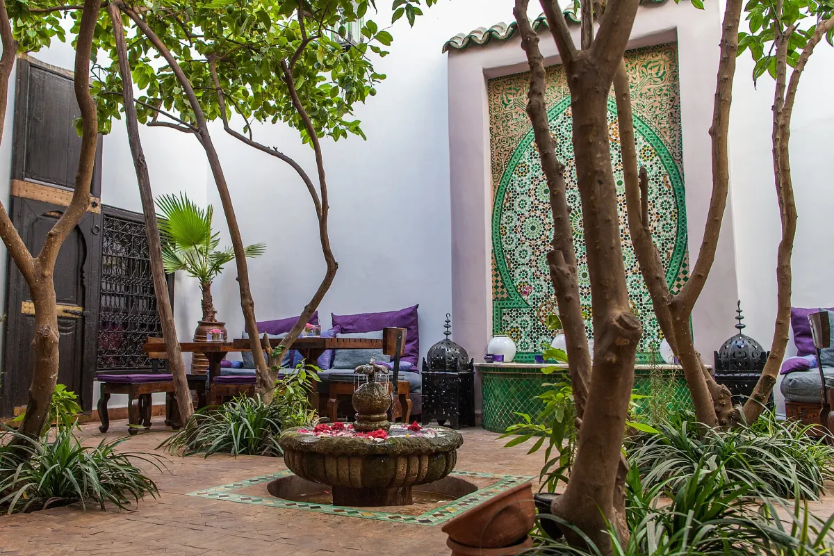 Cour de riad ombragée : fontaine de pierre couverte de pétales de rose, panneau d’arc en zellige vert et blanc, coussins violets le long du mur et plantes en pots sur le sol de brique