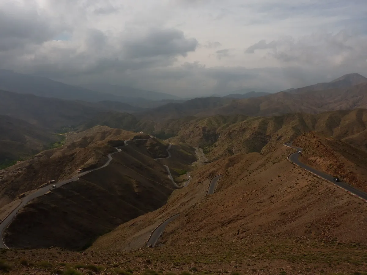 Panorama pris en surplomb sur les lacets du Tizi n’Tichka, déroulés en courbes serrées sur des versants pelés, sous un ciel d’orage et des rideaux de pluie au loin