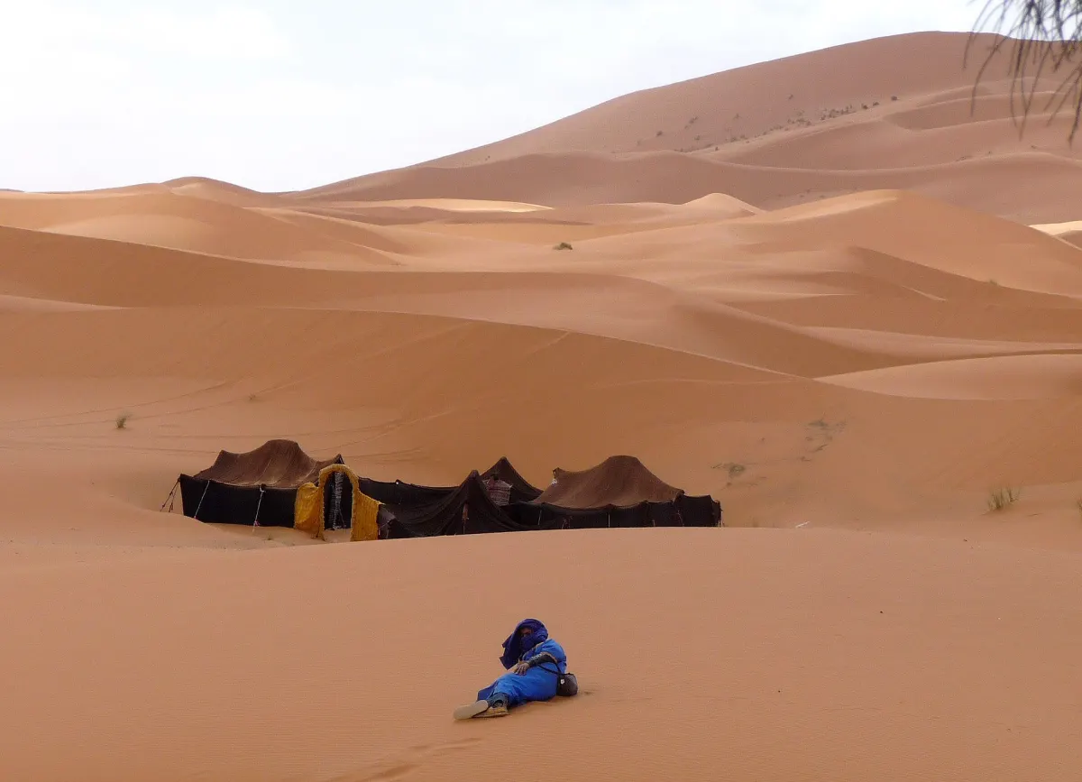 Sous un ciel blanc, des tentes nomades — dont une à l'ouverture frangée de jaune — plantées au pied des dunes, et un homme vêtu de bleu allongé sur le flanc au premier plan